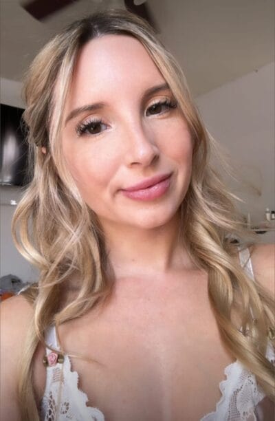 Close-up portrait of a smiling woman with blonde wavy hair wearing a white lace top (selfie-style).