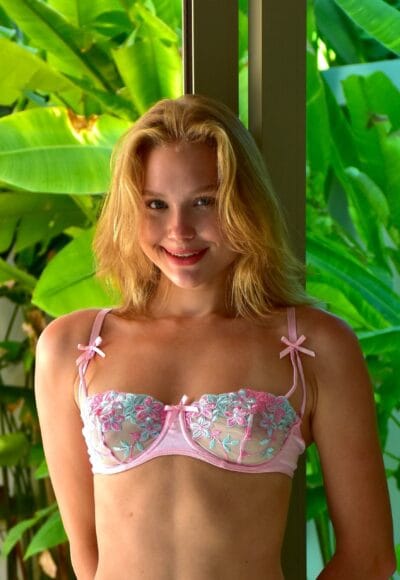 Young woman smiling, wearing a pink lace bra, standing indoors near a window with tropical green plants outside