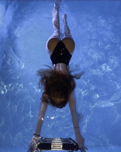 Underwater view of a person in a black bikini reaching for a pool ladder, hair floating in the water.
