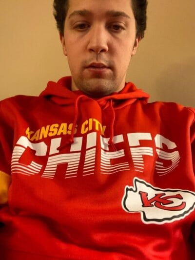 Close-up of a person wearing a red Kansas City Chiefs hoodie, looking downward, neutral expression, beige wall background.