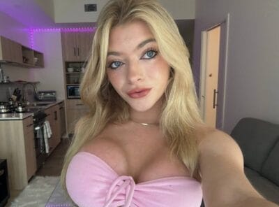 Portrait of a young woman with long blonde hair in a pink off-the-shoulder top, posing in a modern kitchen/living area with purple LED lights behind her.