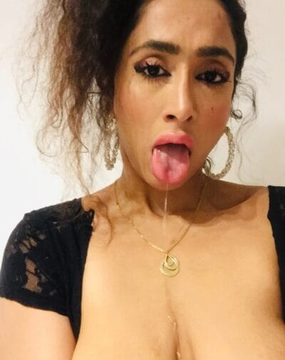 Close-up of a person sticking out their tongue, with a drool string, wearing a black lace top and gold hoop earrings with a pendant necklace.