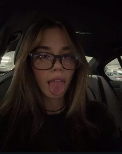 Person in a car at night making a playful face with tongue out, wearing glasses and long hair visible in dim lighting