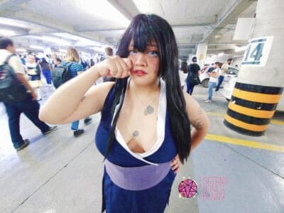 Cosplayer in a dark blue dress with white trim and long black wig, posing with a fist near her face in a busy parking garage.