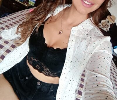 Selfie of a person wearing a white dotted shirt open over a black lace bralette, with a pendant necklace, seated on a bed with a small teddy bear in the background.
