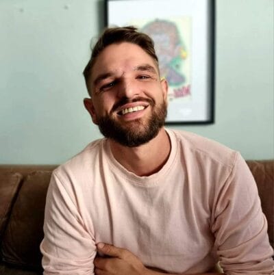 Smiling bearded man in a pale pink sweater sits on a brown couch with a framed poster on the wall behind him.