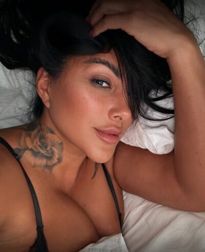 Close-up of a woman lying on a bed with dark hair swept over one eye, hand resting on her head, wearing a black strappy top and a floral neck tattoo visible on her shoulder/neck area.