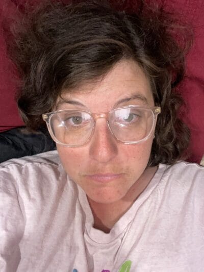 Close-up selfie of a person lying on a red pillow, wearing clear-framed glasses and a light-colored shirt.
