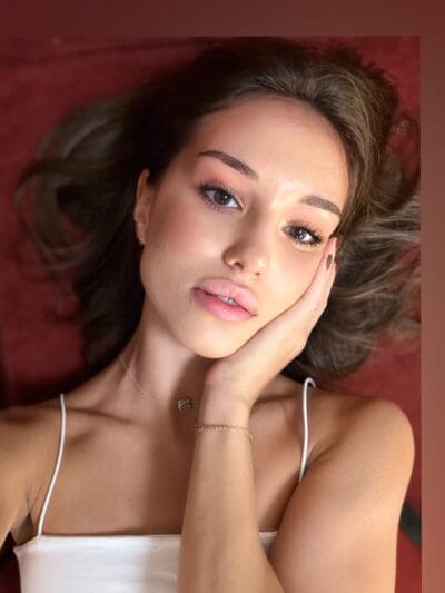 Close-up of a young woman with brown hair lying on a red background, head resting on her hand, wearing a white spaghetti-strap top.