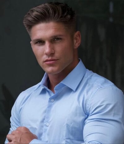 Portrait of a man with short brown hair wearing a light blue dress shirt, looking at the camera with a neutral expression on a dark background.
