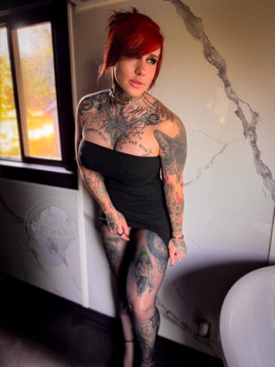 Tattooed woman with bright red hair in a black strapless dress stands indoors near a cracked marble wall by a window with autumn foliage visible outside, showcasing full-body tattoos.