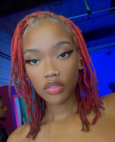 Close-up portrait of a young woman with red dreadlocks, winged eyeliner, and a nose ring, under blue lighting.