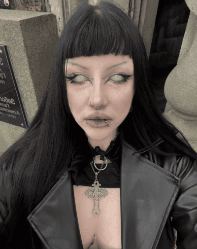 Person with long black hair and blunt bangs, gothic makeup, and multiple facial piercings wearing a black leather jacket and a large ornate cross pendant outside a stone building.