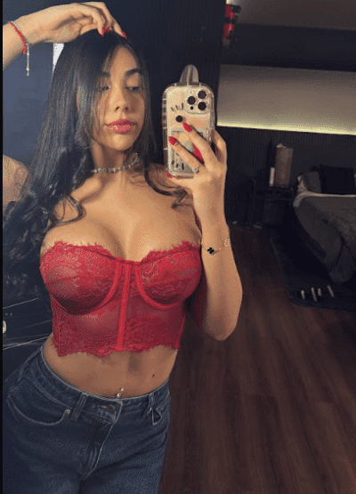 Woman in a red lace bralette posing for a mirror selfie in a bedroom, holding a phone with a clear case and long dark hair falling over her shoulders.