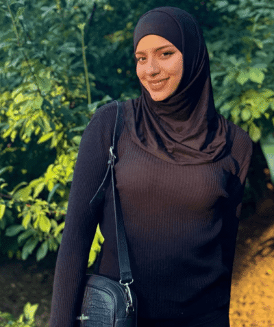 Smiling woman in a black hijab and sweater outdoors, carrying a black crossbody bag in a sunny garden setting.