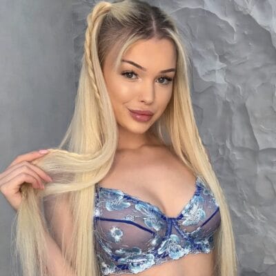 Woman with long blonde hair, wearing a blue floral lace bralette, holding her hair and smiling softly at the camera.