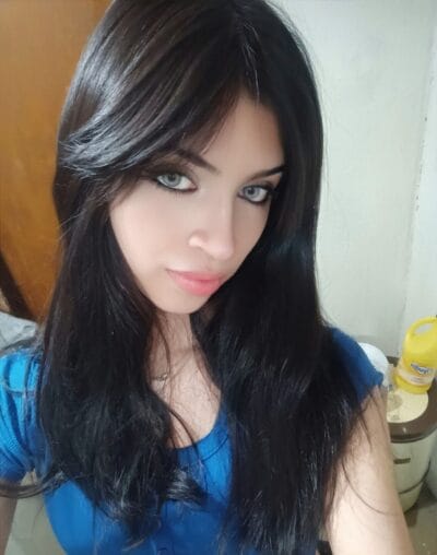 Close-up of a young woman with long dark hair and blue eyes, wearing a blue top, looking at the camera outdoors/indoors.
